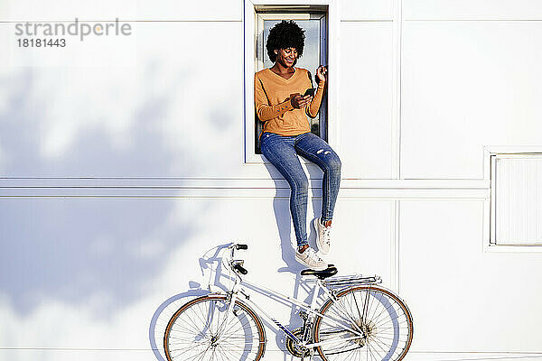 Lächelnde Frau mit Fahrrad und Mobiltelefon auf der Fensterbank