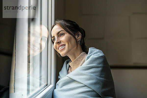 Nachdenkliche junge Frau  in eine Decke gehüllt  steht am Fenster