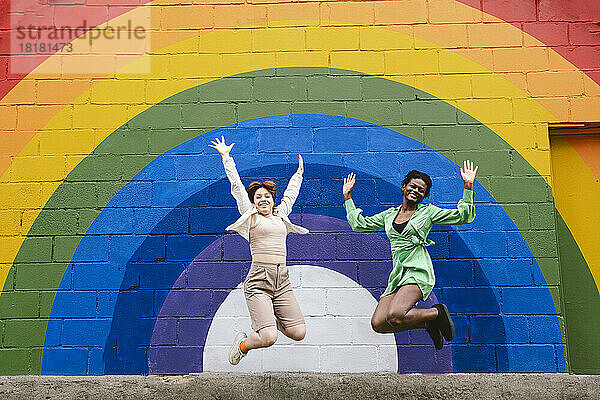Lächelnde junge Frau mit Freundin springt vor einer an die Wand gemalten Regenbogenfahne