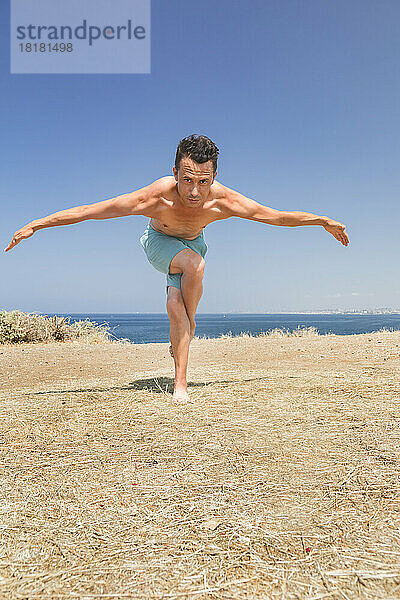 Hemdloser Mann praktiziert an einem sonnigen Tag Adler-Pose-Yoga auf einem Feld vor dem Meer