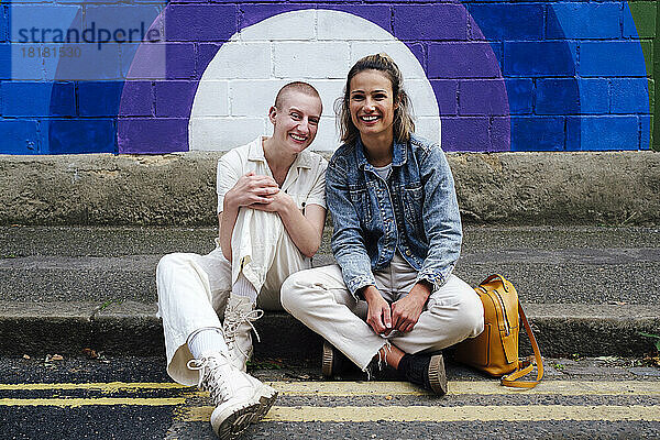 Glückliche junge Frau und nicht-binäre Person sitzen auf der Straße