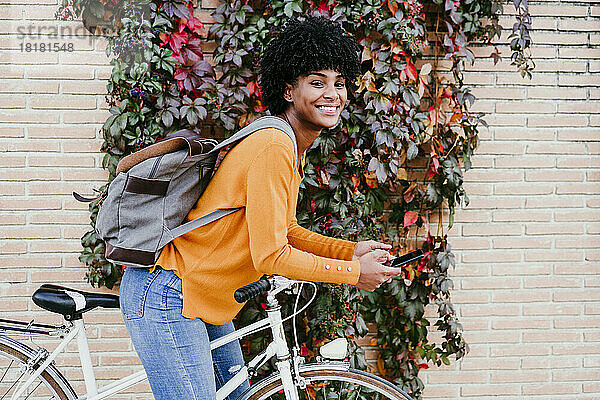 Lächelnde junge Frau mit Rucksack und Smartphone auf dem Fahrrad
