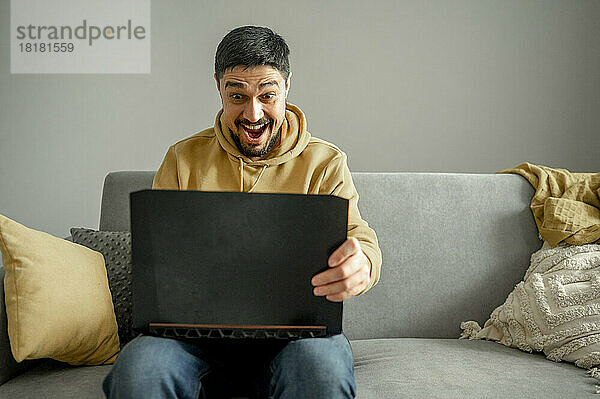 Überraschter Mann benutzt Laptop  der zu Hause auf dem Sofa sitzt
