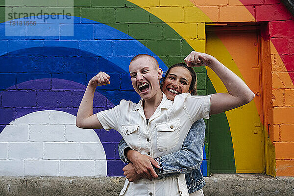 Glückliche lesbische Frau umarmt nicht-binäre Person und lässt vor der Regenbogenwand Muskeln spielen