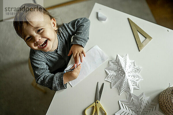 Fröhlicher Junge sitzt mit Papierschneeflocken am Tisch