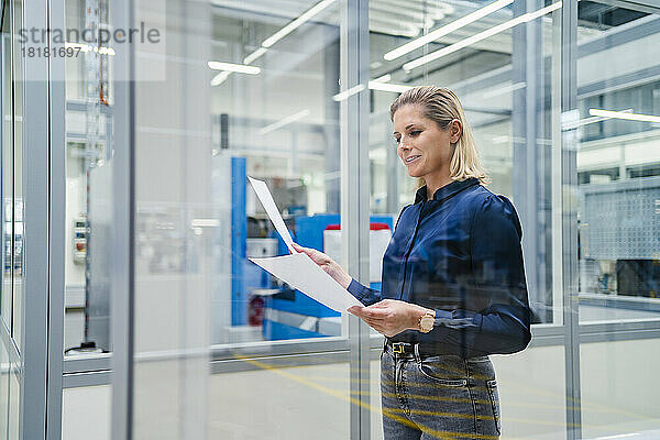 Lächelnde Geschäftsfrau überprüft Dokumente in der Fabrik  gesehen durch Glas