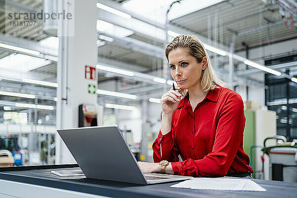 Konzentrierte Geschäftsfrau mit der Hand am Kinn und Blick auf den Laptop in der Fabrik