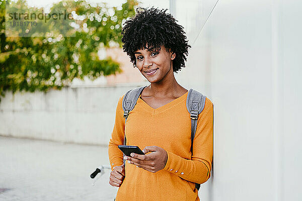 Lächelnde junge Frau mit Mobiltelefon an der Wand