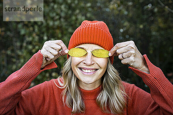 Lächelnde reife Frau bedeckt Augen mit Herbstblättern
