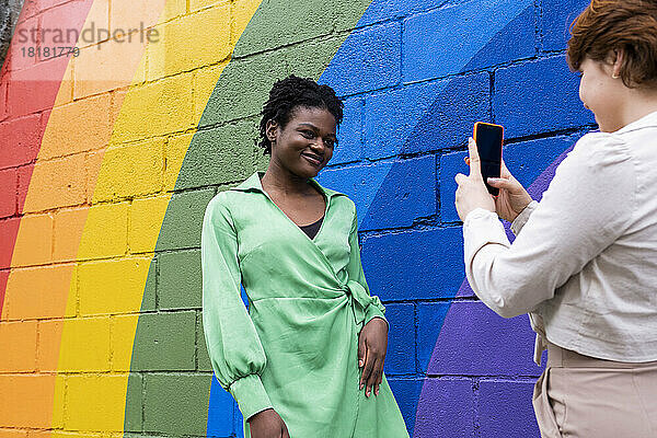 Lächelnde Frau fotografiert Freundin  die neben einer an die Wand gemalten Regenbogenfahne steht