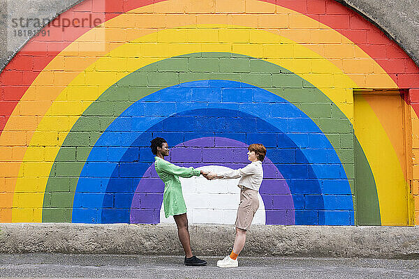 Frau hält die Hand ihrer Freundin vor einer an die Wand gemalten Regenbogenfahne