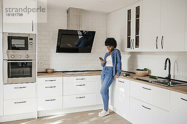 Junge Frau benutzt Smartphone und lehnt sich zu Hause an die Küchentheke
