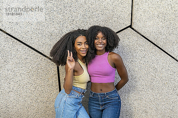 Smiling woman gesturing peace sign standing with friend in front of wall