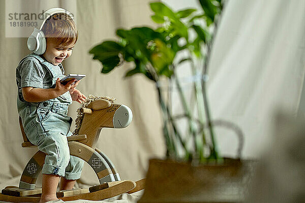 Fröhlicher Junge mit kabellosen Kopfhörern und Smartphone auf Schaukelpferd