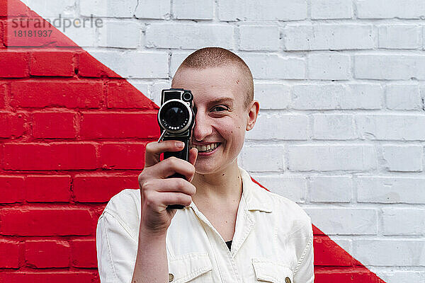 Nicht-binäre Person bedeckt Auge mit Videokamera vor zweifarbiger Wand