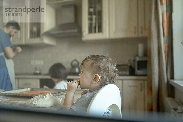 Durch das Fenster gesehener kleiner Junge  der in der Küche Pizzateig isst