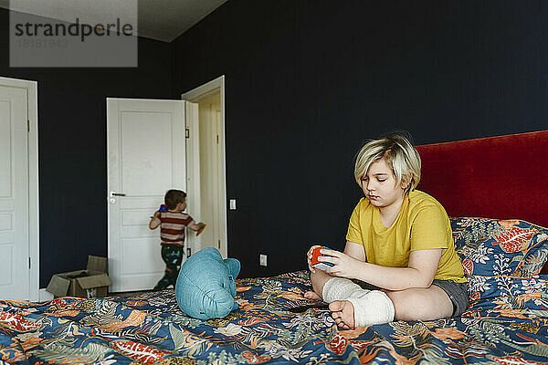 Junge mit Gips am Bein spielt mit Puzzle-Würfel und sitzt zu Hause im Bett