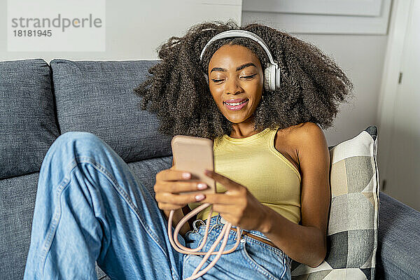 Lächelnde Frau benutzt Smartphone und hört zu Hause Musik über kabellose Kopfhörer