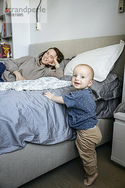 Netter Junge  der neben dem Bett steht  mit der Mutter im Hintergrund