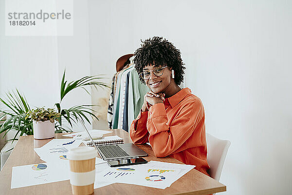 Glückliche Geschäftsfrau sitzt mit der Hand am Kinn am Schreibtisch im Heimbüro