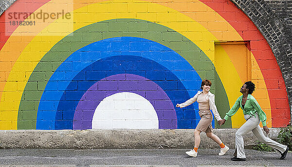 Lächelndes lesbisches Paar hält sich an den Händen und läuft auf dem Fußweg vor einer regenbogenfarbenen Wand