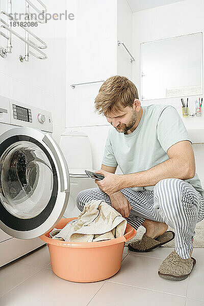 Mann benutzt Smartphone neben Waschmaschine im Badezimmer zu Hause