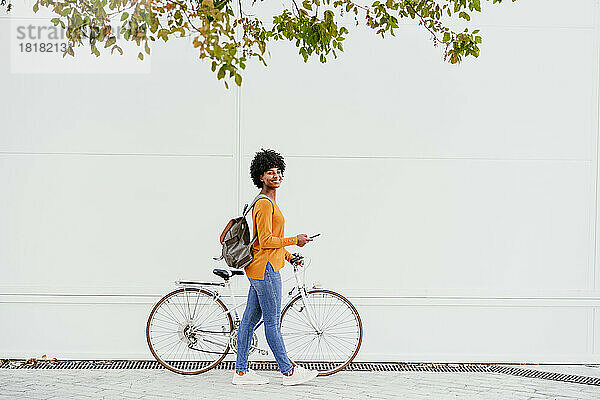 Lächelnde Frau mit Rucksack und Smartphone  die ihr Fahrrad an der Wand vorbeiführt