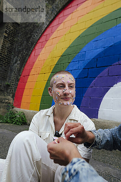 Nicht-binäre Person mit Aufkleber auf dem Gesicht sitzt neben einem Freund vor einer bunten Wand