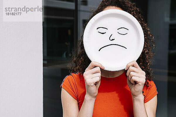 Junge Frau hält trauriges Emoticon-Teller über Gesicht