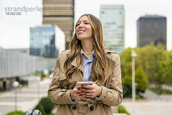 Lächelnde Geschäftsfrau hält Mobiltelefon vor Gebäuden