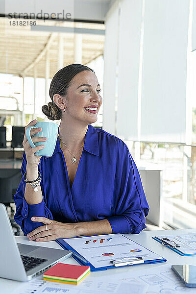 Lächelnde junge Geschäftsfrau mit Kaffeetasse am Schreibtisch im Büro