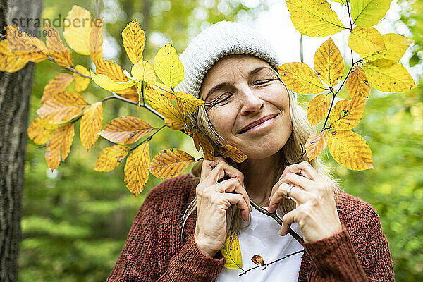 Lächelnde Frau mit geschlossenen Augen hält herbstlichen Pflanzenstamm im Wald