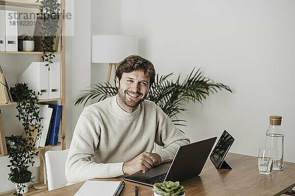 Lächelnder Geschäftsmann mit Laptop auf dem Schreibtisch im Büro