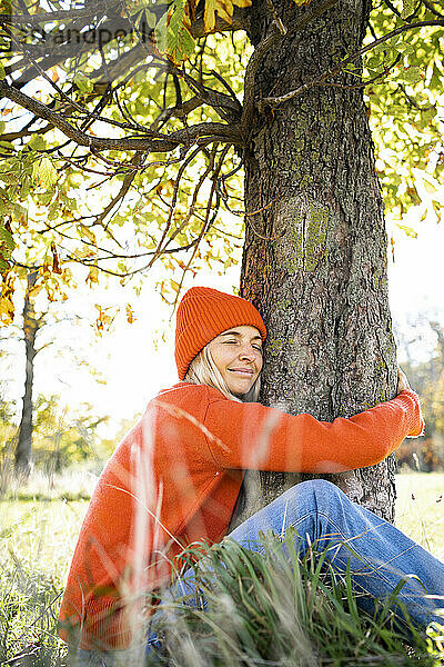 Lächelnde Frau umarmt Baumstamm am Wochenende im Herbst