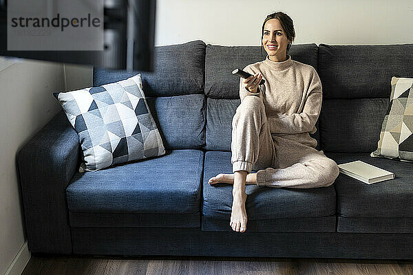 Smiling woman holding remote control sitting on sofa at home