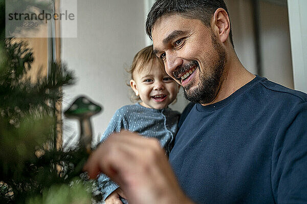 Glücklicher Vater trägt seinen Sohn und schmückt den Weihnachtsbaum zu Hause