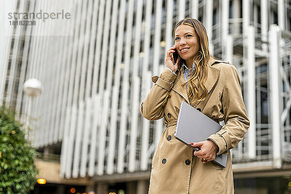 Lächelnde Geschäftsfrau mit Laptop und Telefon vor dem Gebäude