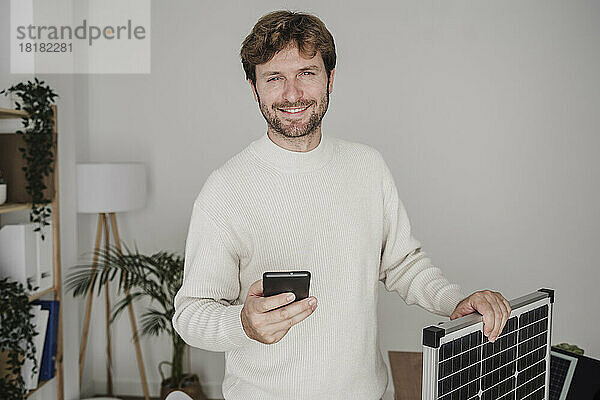 Lächelnder Geschäftsmann mit Smartphone und Solarpanel im Büro