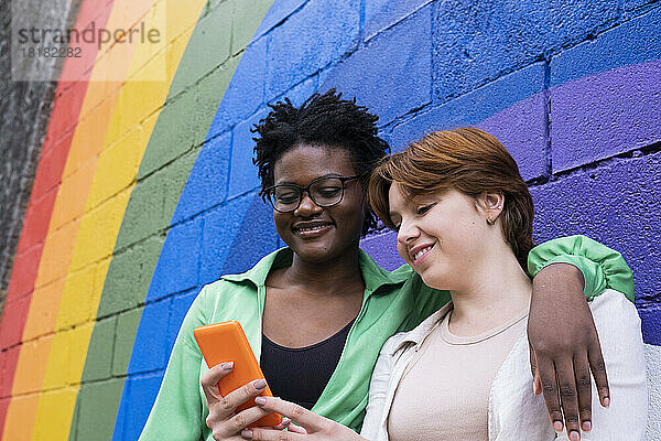 Lächelnde Frau teilt Smartphone mit Freund vor regenbogenfarbener Wand