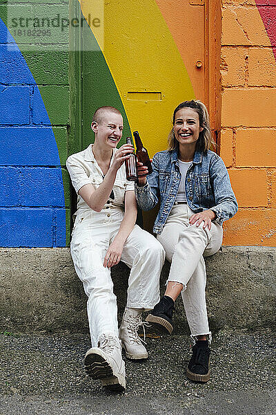 Glückliche junge Frau prostet Bier mit nicht-binärer Freundin zu  die vor einer bunten Wand sitzt