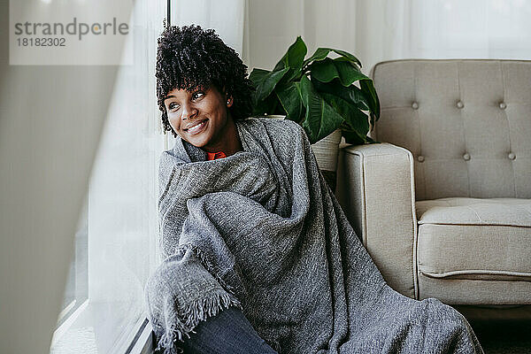 Nachdenkliche junge Frau  in eine graue Decke gehüllt  sitzt zu Hause am Fenster