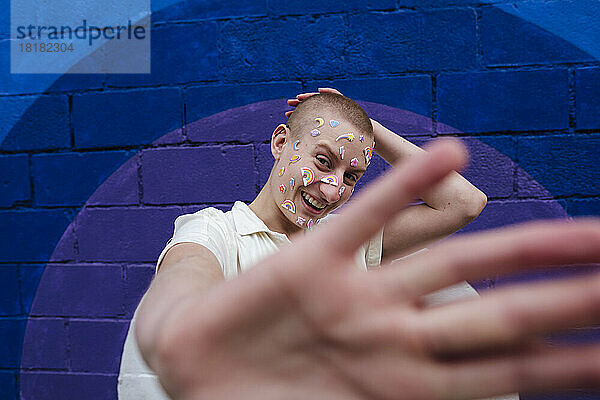 Fröhliche nicht-binäre Person mit Aufklebern im Gesicht  die eine Stopp-Geste vor einer bunten Wand zeigen