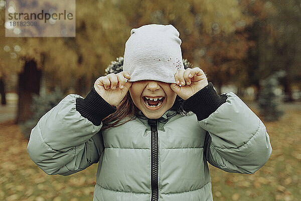 Fröhliches Mädchen bedeckt Gesicht mit Strickmütze im Herbstpark