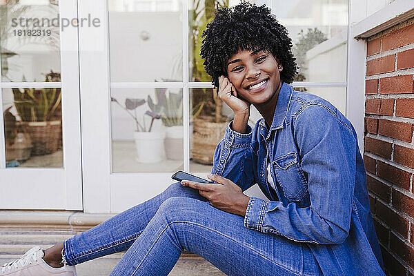 Glückliche junge Frau mit Smartphone sitzt im Türrahmen
