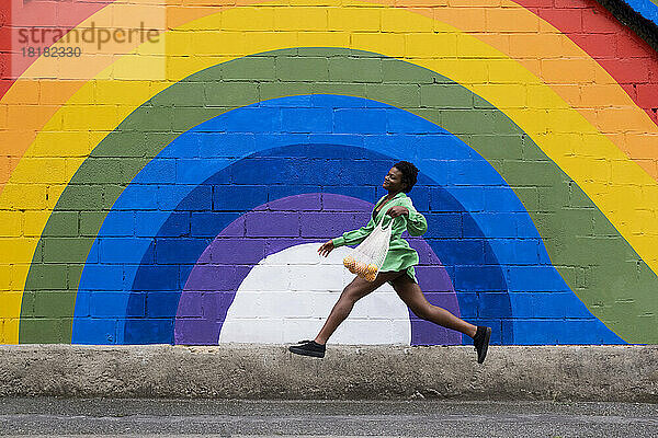 Lächelnde junge Frau mit Netzbeutel springt vor einer an die Wand gemalten Regenbogenfahne