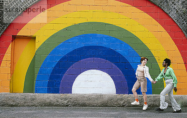 Glückliches lesbisches Paar hält sich an den Händen und geht auf dem Fußweg vor einer regenbogenfarbenen Wand