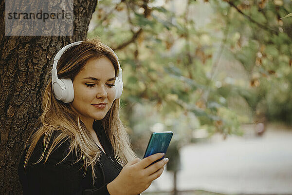 Lächelnde Frau  die im Park Musik über kabellose Kopfhörer hört und ihr Smartphone benutzt