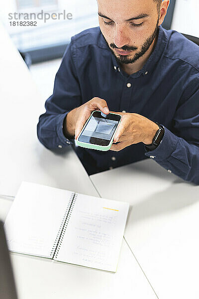 Junger Geschäftsmann fotografiert mit Smartphone am Schreibtisch