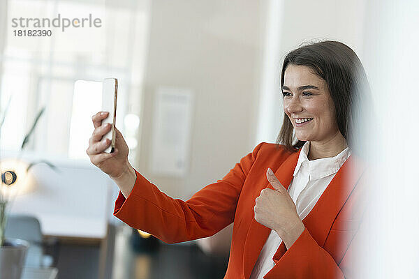 Glückliche Geschäftsfrau zeigt Daumen hoch bei Videoanruf über Smartphone im Büro