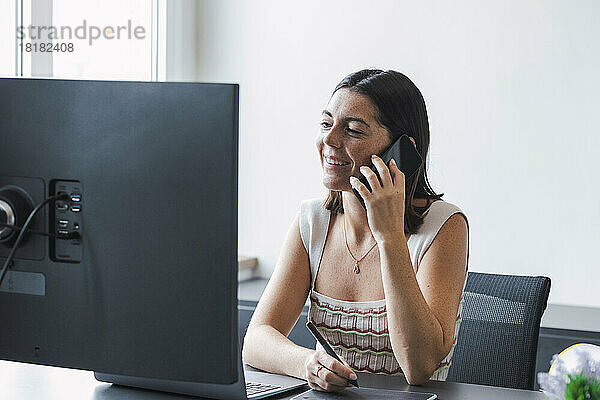 Lächelnde Geschäftsfrau  die am Schreibtisch im Büro sitzt und über ihr Smartphone spricht